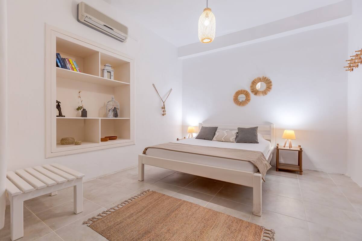 Un dormitorio con cama, mesitas de noche, un banco, una estantería con libros y adornos en la pared.