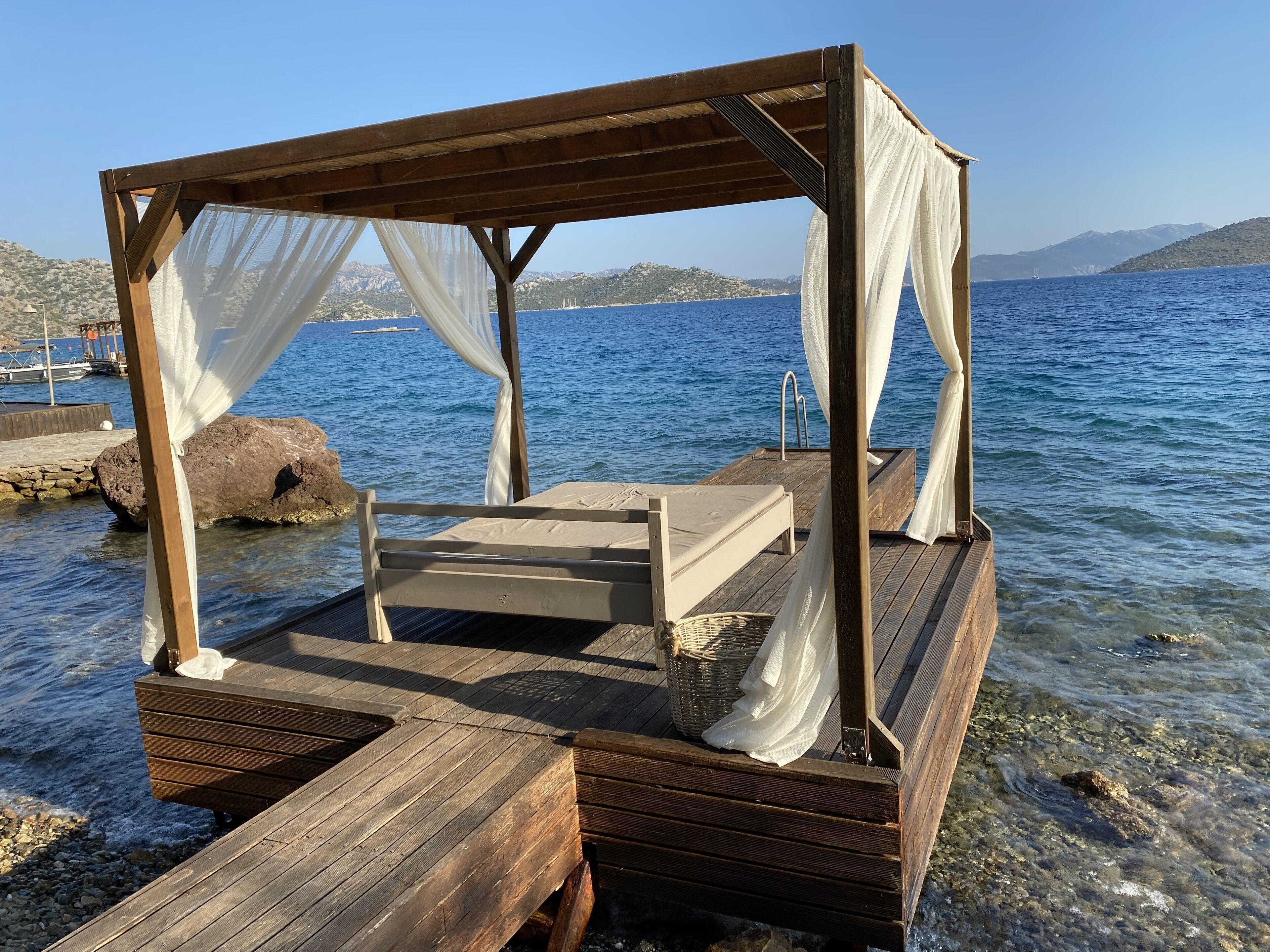 Terraza de madera con una cama y dosel, con vista a un cuerpo de agua con barcos y colinas distantes.