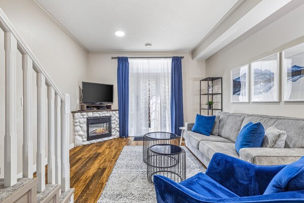 Ein Wohnzimmer mit Kamin aus Stein, einem grauen Sofa mit blauen Kissen, einem Fernseher und einer Treppe mit weißen Geländern.