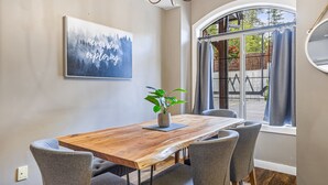 Een eetruimte met een houten tafel, zes grijze stoelen, een potplant en een groot raam met gordijnen.