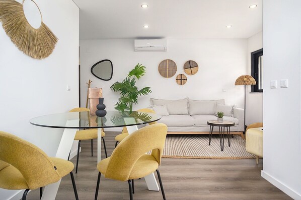 A modern living room with a glass dining table, yellow chairs, a white sofa, and a small side table with a plant.