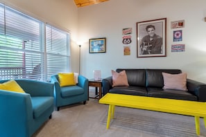 Un salon avec un canapé bleu, un canapé noir, un banc jaune et un tableau encadré accroché au mur.