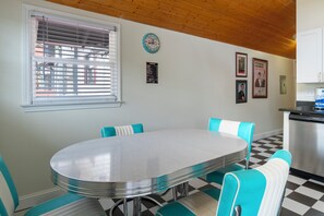 Un coin repas avec une table ronde et des chaises de couleur turquoise, une fenêtre avec des stores, une horloge et des tableaux encadrés accrochés au mur.