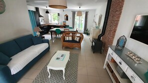Una sala de estar con sofá, una mesa de centro, un sillón de mimbre, un comedor con mesa y sillas, una cocina con estufa, y un televisor.