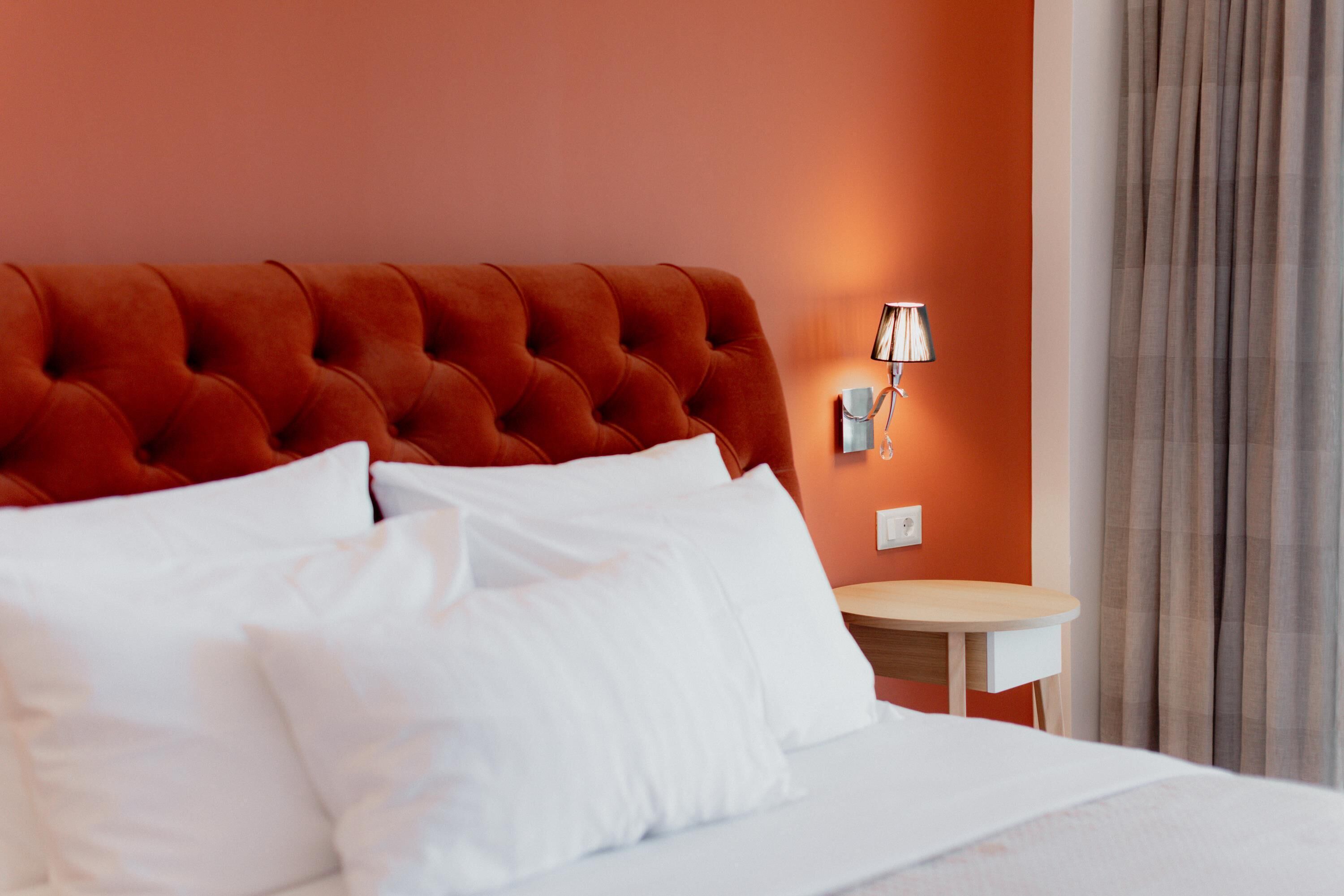 Une chambre d’hôtel avec une tête de lit capitonnée, du linge de lit blanc, une table de chevet avec une lampe et une vue sur un rideau.