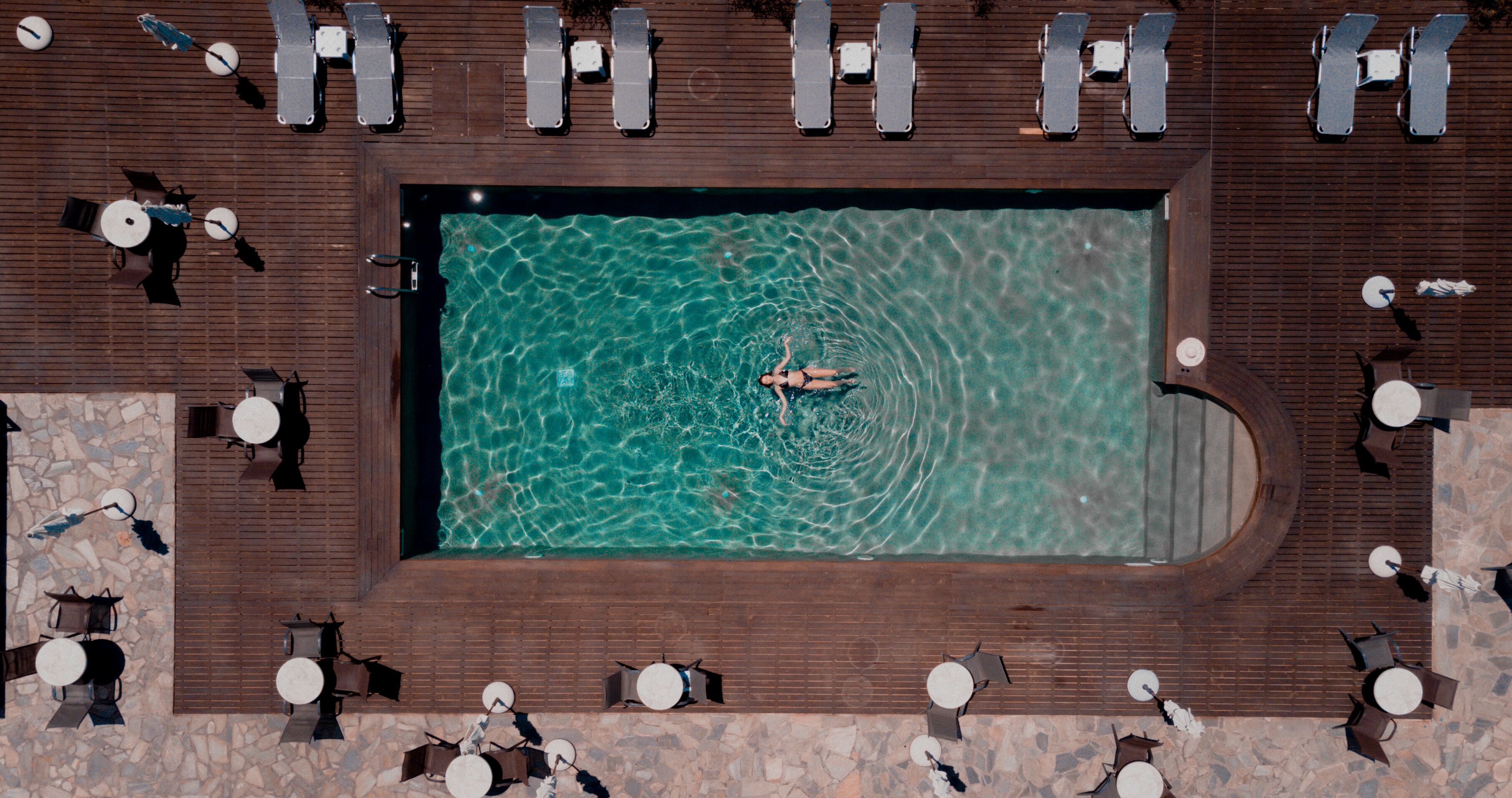 An aerial view of a rectangular swimming pool surrounded by lounge chairs and tables, with a person swimming in the pool.
