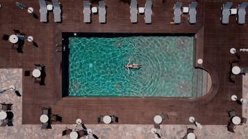 An aerial view of a rectangular swimming pool surrounded by lounge chairs and tables, with a person swimming in the pool.