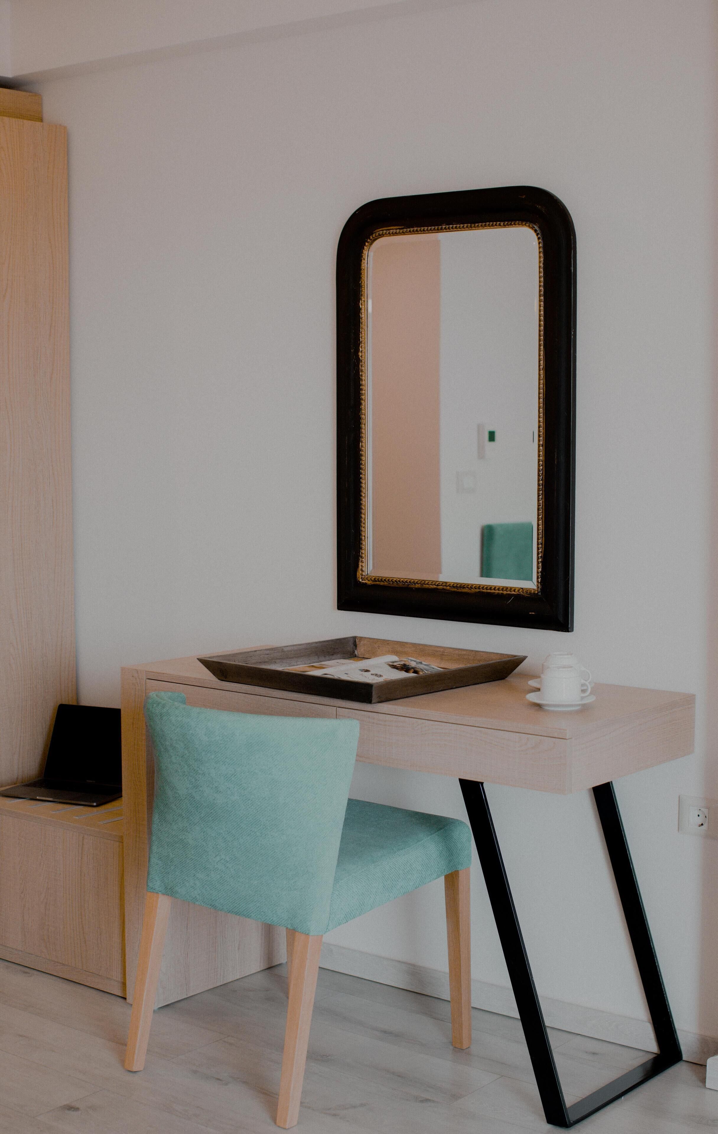 Une petite pièce moderne, dotée d’une coiffeuse en bois, d’un miroir, d’un ordinateur portable et d’un fauteuil de couleur turquoise.