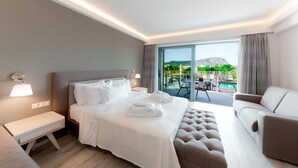 A modern hotel room with a large bed, a sofa, a bench, and a view of a pool and mountains. - Palacio Del Sol Luxury Apartments (Nafplio)