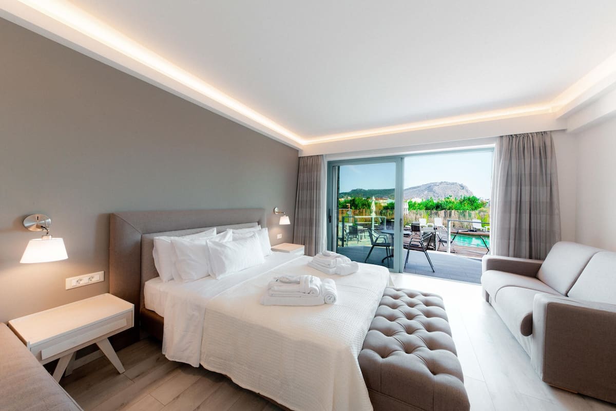 Habitación de hotel moderna con una cama grande, un sofá, un banco y vistas a una piscina y montañas.