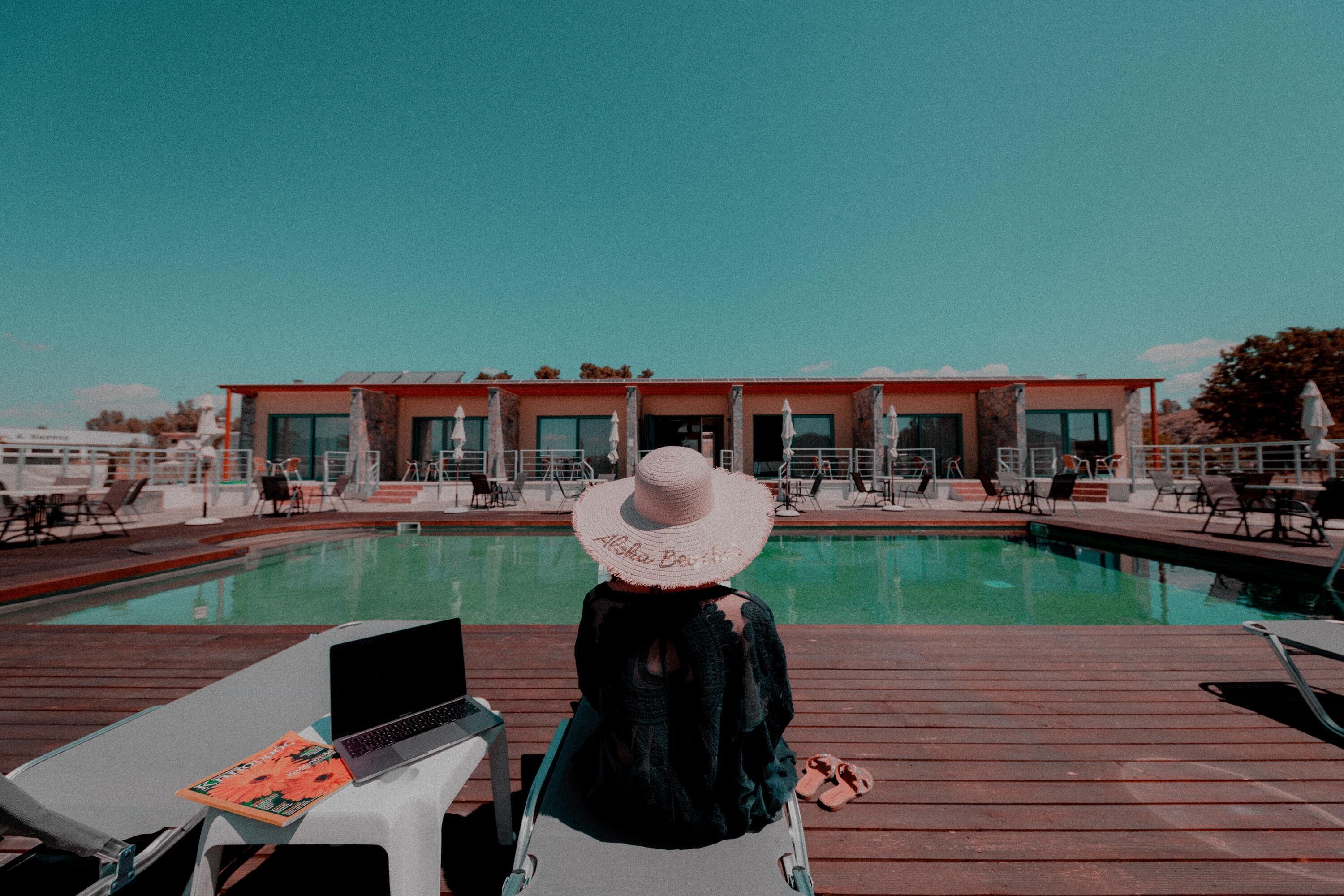 A person sitting by a pool with a laptop, overlooking a building with a sign that reads "Alaska Beach."