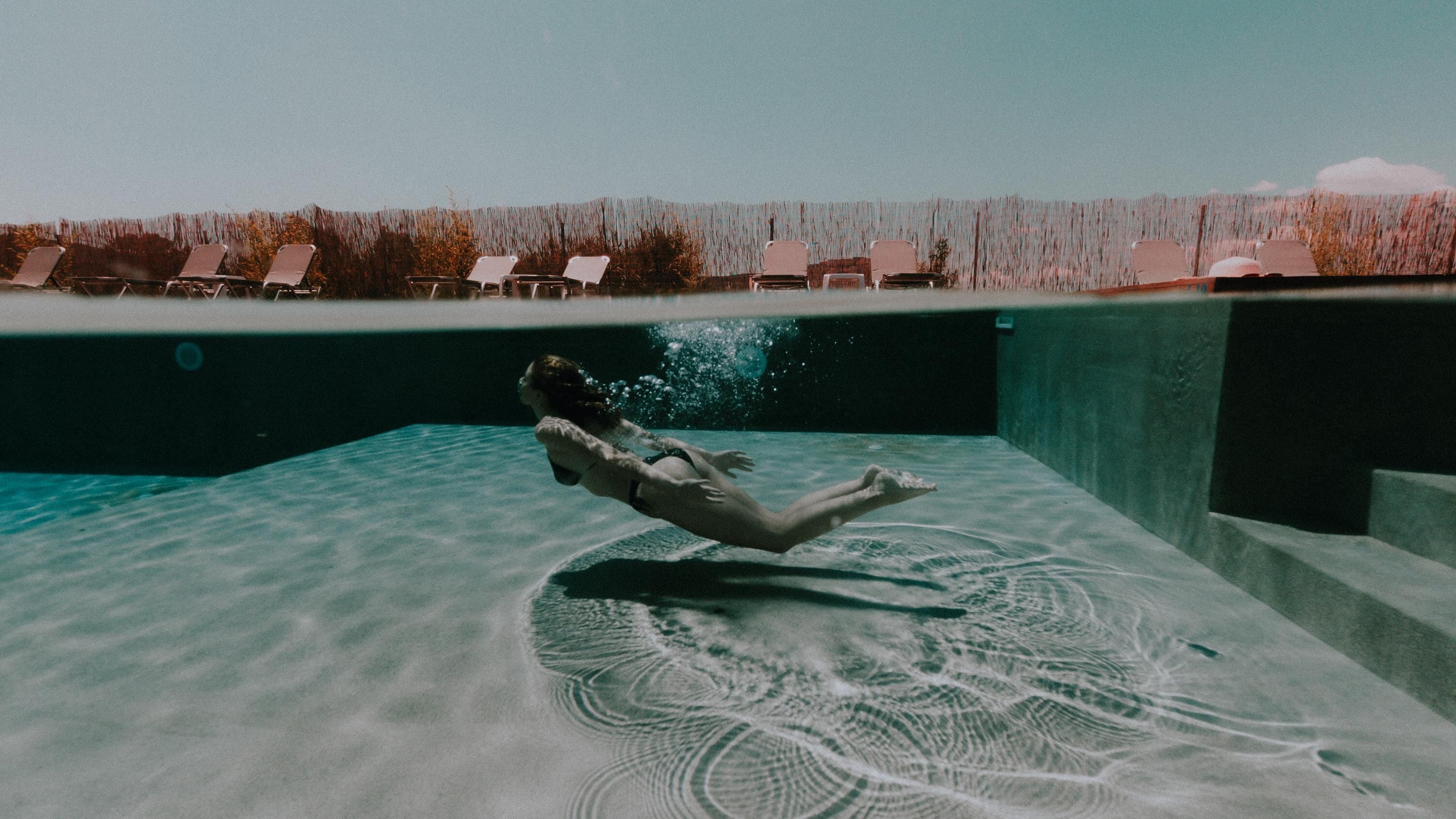 A person swimming in a pool with a view of lounge chairs and a fence in the background.