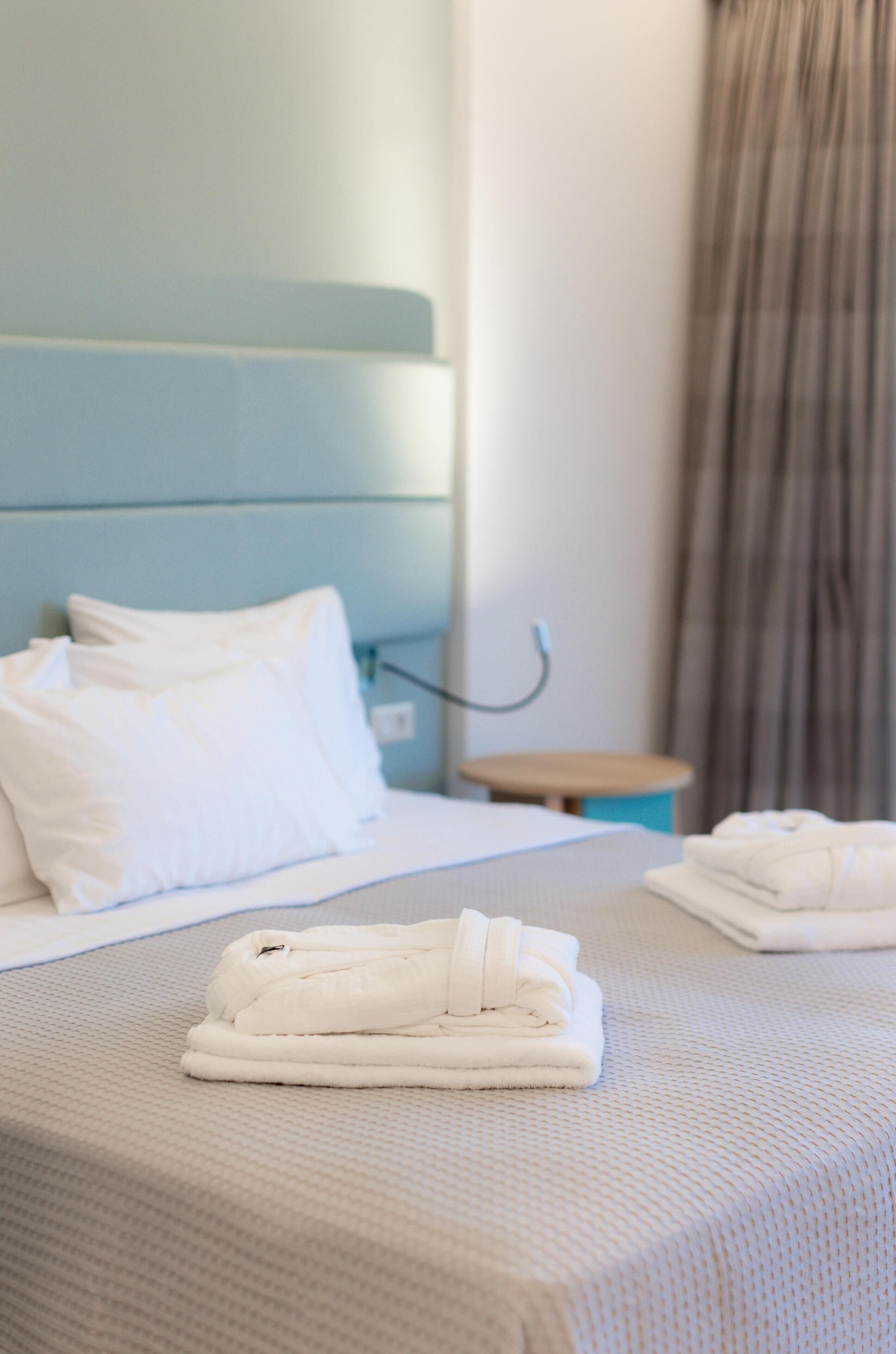 Una cama bien hecha con ropa blanca y almohadas, una mesita de noche con lámpara, y una ventana con cortinas.