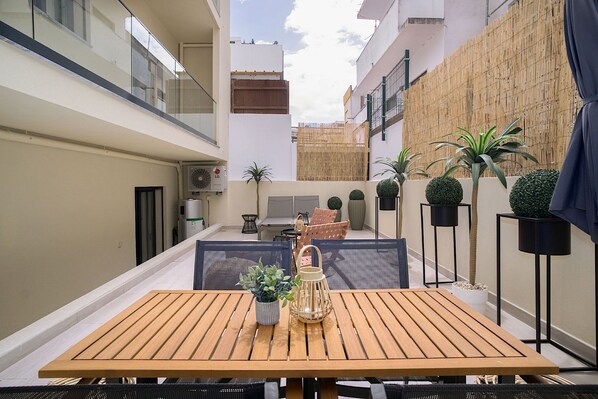 Un patio con mesa de madera, sillas, plantas en macetas y un pequeño espacio de estar.