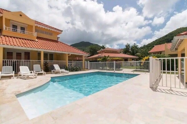A swimming pool with white lounge chairs, surrounded by a white fence, and buildings with red-tiled roofs in the background.