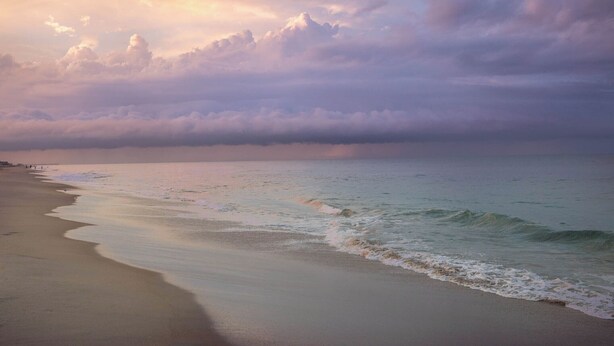 A beach with waves gently lapping onto the shore, a pier in the distance, and a cloudy sky.