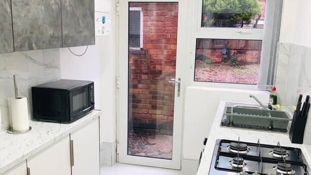 A compact kitchen with white cabinets, a black microwave, a gas stove, and a window with a view of a brick wall and greenery outside. - 4 Bedroom Lovely Home in Loughborough Town & Uni (Loughborough)