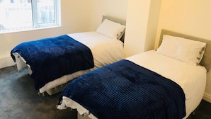 A room with two beds, each with a white mattress and blue bedspread, and a window providing a view of buildings outside. - 4 Bedroom Lovely Home in Loughborough Town & Uni (Loughborough)