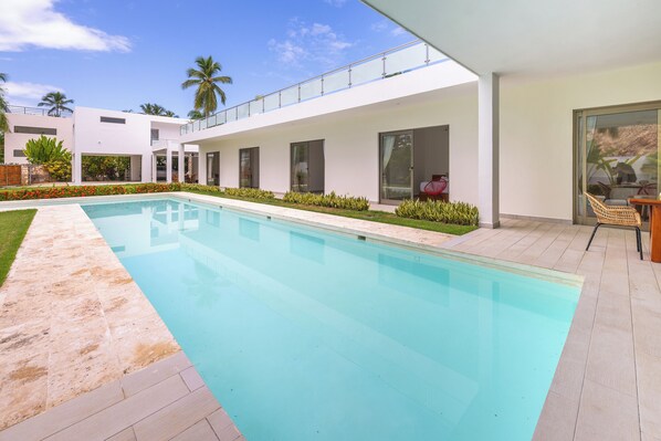 A modern poolside area with a clear pool, a covered seating area, and a white building with large windows.