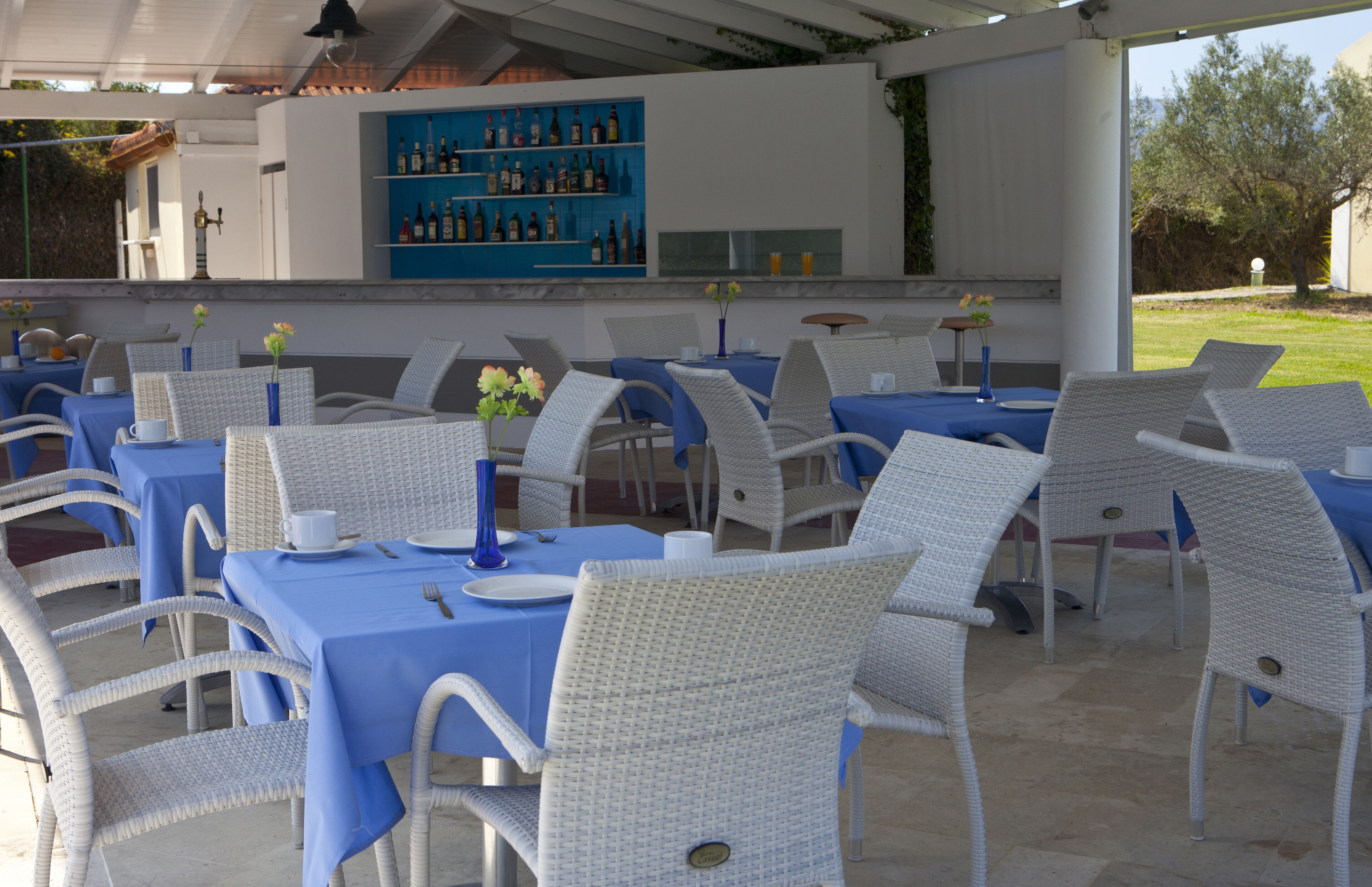 Un espace extérieur aménagé pour les repas, avec des chaises en osier blanc et des nappes bleues, un bar doté d’étagères bleues, et une vue sur le jardin.