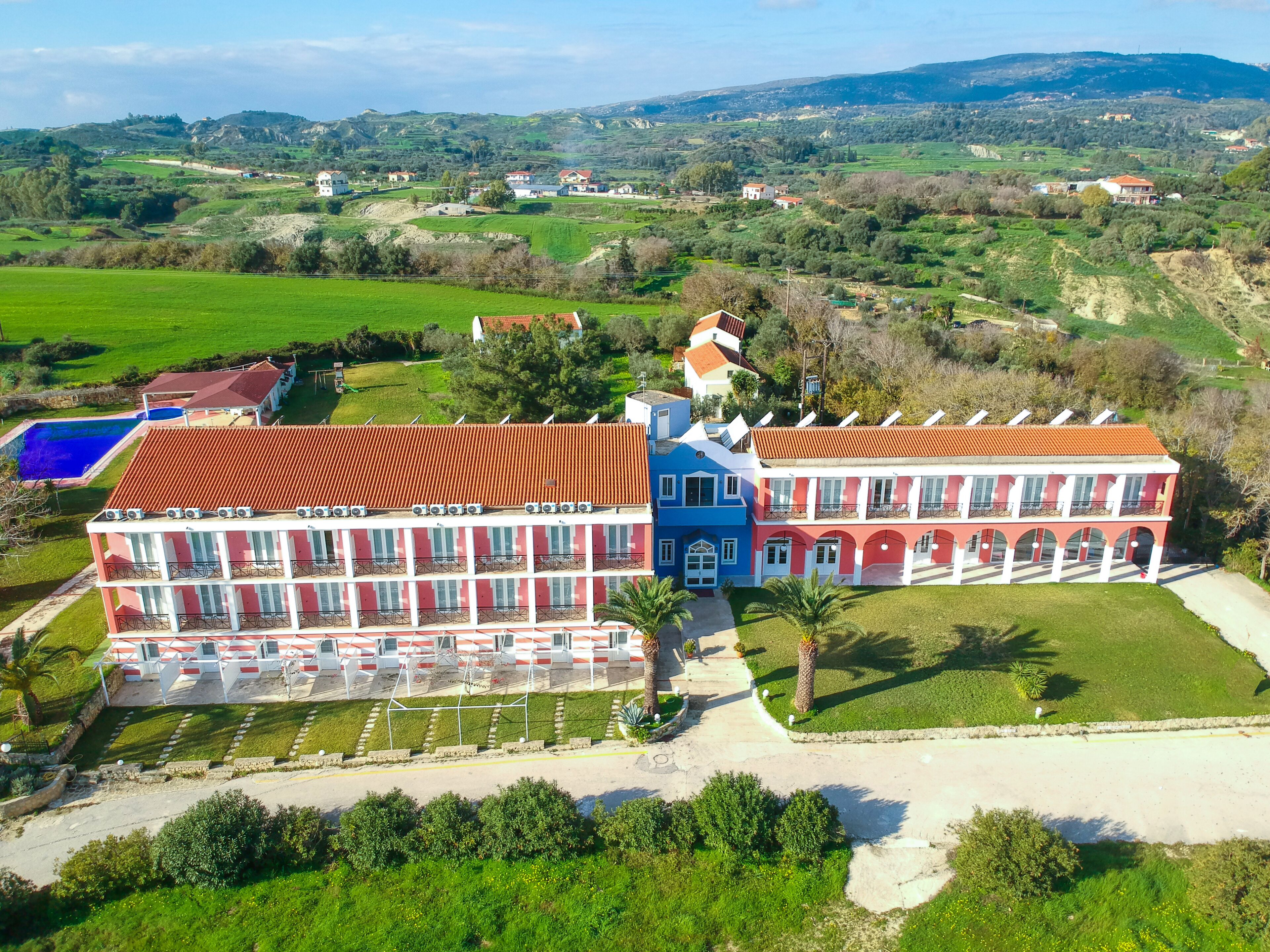 Un grand immeuble à étages, doté d’une façade rouge et bleue, entouré de vastes champs verdoyants et surplombé d’un ciel dégagé.