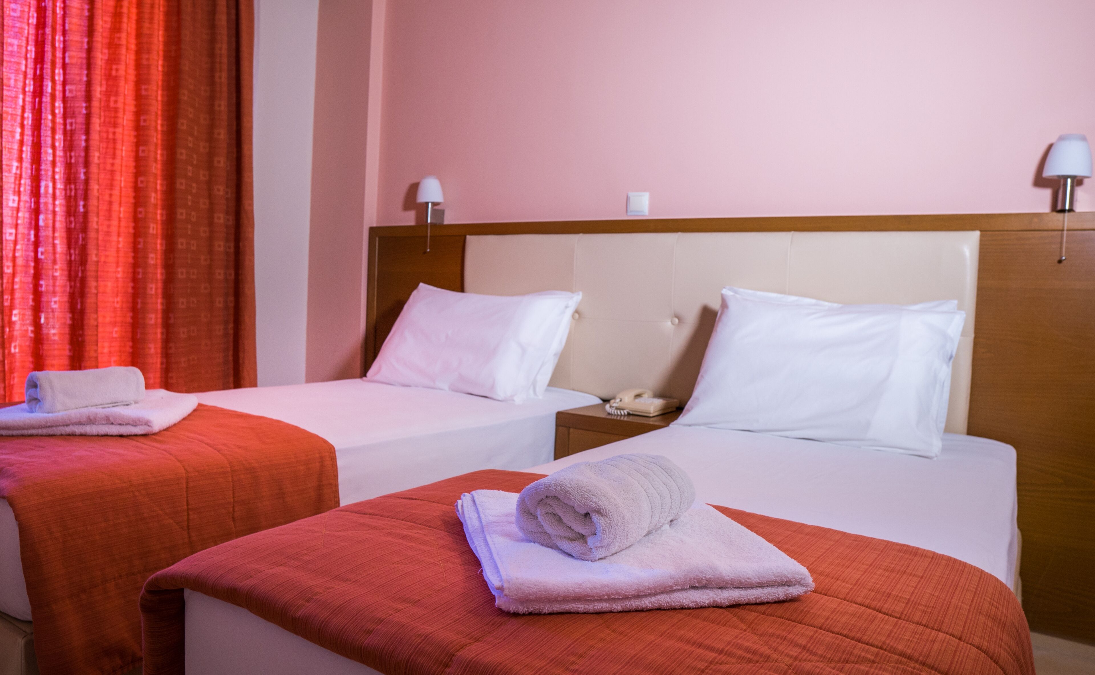 A hotel room with two beds, each with a white bedspread and pillows, a wooden headboard, a nightstand with a telephone, and a red curtain.