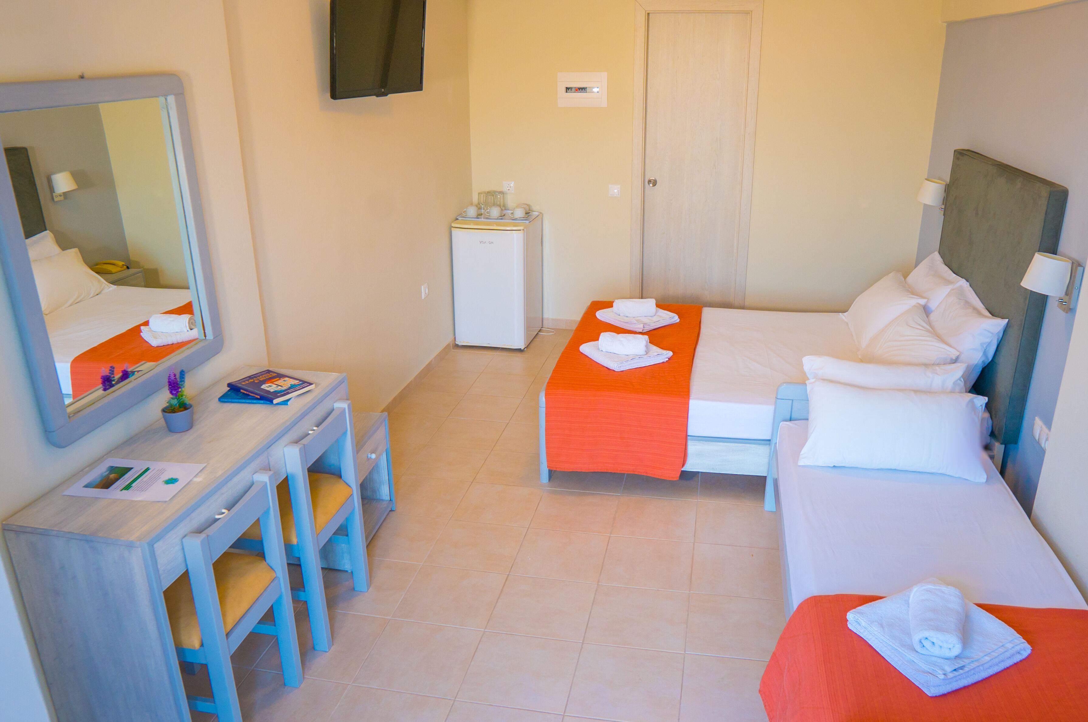 A hotel room with a bed, a desk with chairs, a small refrigerator, and a mirror.