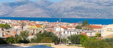 A coastal town with red-roofed buildings, a clear blue sea, and mountains in the background.