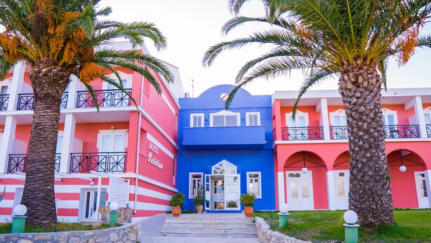 Un hotel con una fachada vibrante, destacando un edificio central azul flanqueado por edificios rosados y rojos. Palmeras bordean el camino que lleva a la entrada.