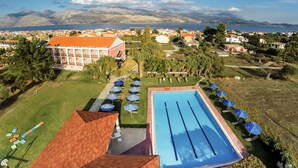 Una piscina con sillones de descanso y sombrillas, rodeada por un edificio con techo rojo y vista a montañas y una masa de agua al fondo.