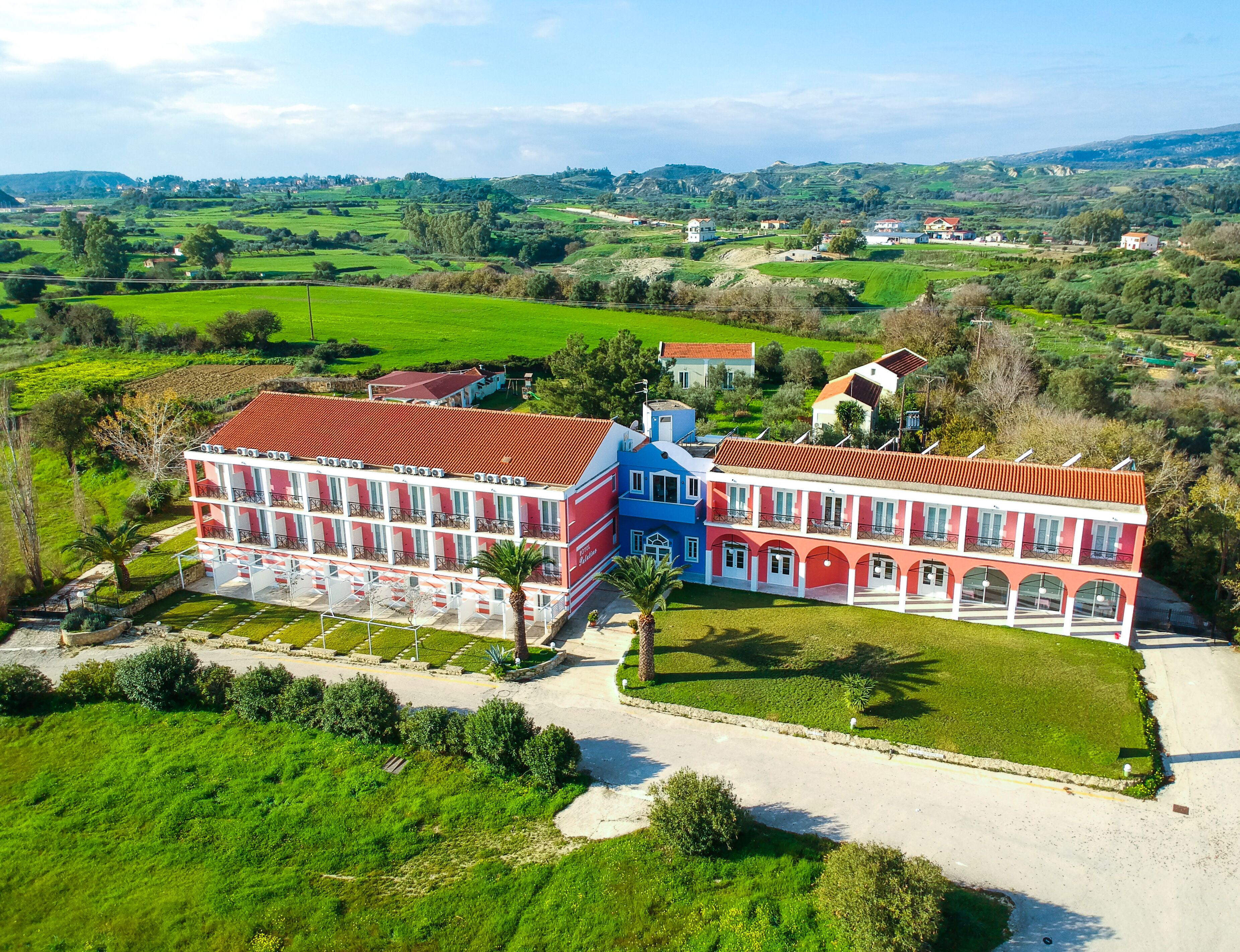 Um edifício grande e com vários andares, com fachada em vermelho e branco, cercado por campos verdes exuberantes e um céu claro.