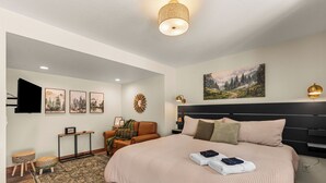 Een slaapkamer met een groot bed, een televisie, een wandlamp, een schilderij en een kleine tafel met lamp.