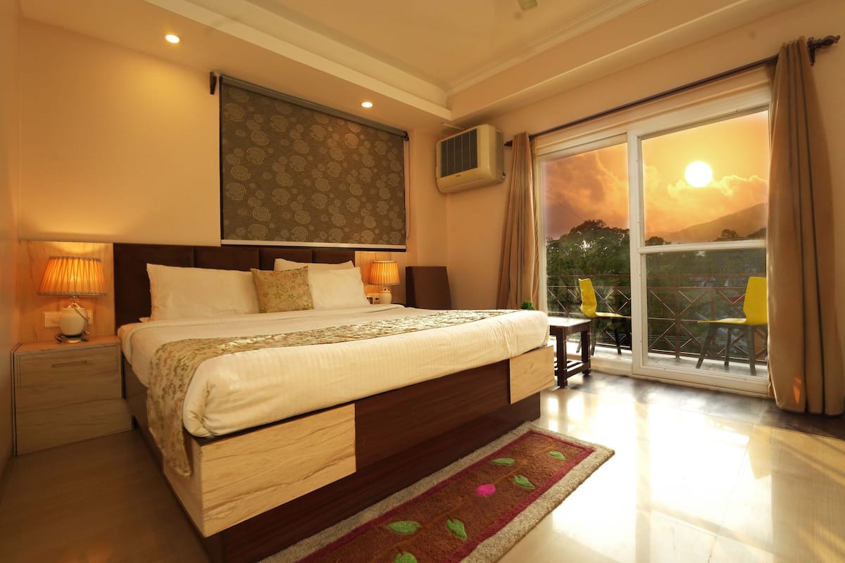 A spacious bedroom with a large bed, a nightstand, a bedside lamp, a window with a view of the sunset, and a sliding glass door leading to an outdoor area.