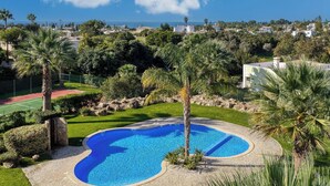 Piscina rodeada de palmeras y una cancha de tenis, con vistas a casas y al mar al fondo.