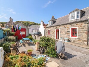 A stone building with a red door and window, a car parked in front, and a garden with plants and a wooden bench. - Dookers' Hoose (Buckie)