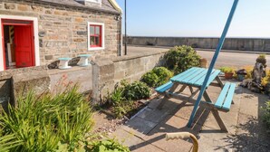 A stone building with a red door and a small garden with a picnic table. - Dookers' Hoose (Buckie)