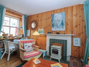 Une pièce chaleureuse aux murs en bois, avec une cheminée, une table à manger et un fauteuil agrémenté d’un coussin coloré.