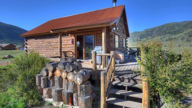 A log cabin with a metal roof, a porch with outdoor seating, and a view of the surrounding mountains. - Rustic Log Cabin - Close to Creede - Picturesque Views (Creede)