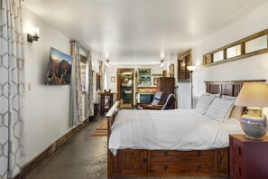 Ein Schlafzimmer mit einem großen Bett, einem Holzkopfende und einem Nachttisch mit Lampe. Im Raum befinden sich eine Bank und ein Sessel, sowie eine kleine Küchenzeile im Hintergrund.