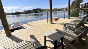 A covered deck with lounge chairs and a table, overlooking a calm lake with a house in the background. - Dockside Daze-Prime Waterfront Loc w/ Amazing View, Firepit, Tiki Bar, Lrg Dock (Moneta)