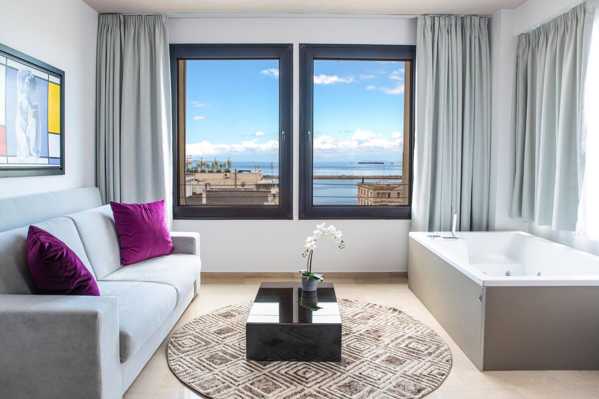 Een moderne hotelkamer met een bank, een bijzettafeltje, een badkuip en grote ramen met uitzicht op zee.