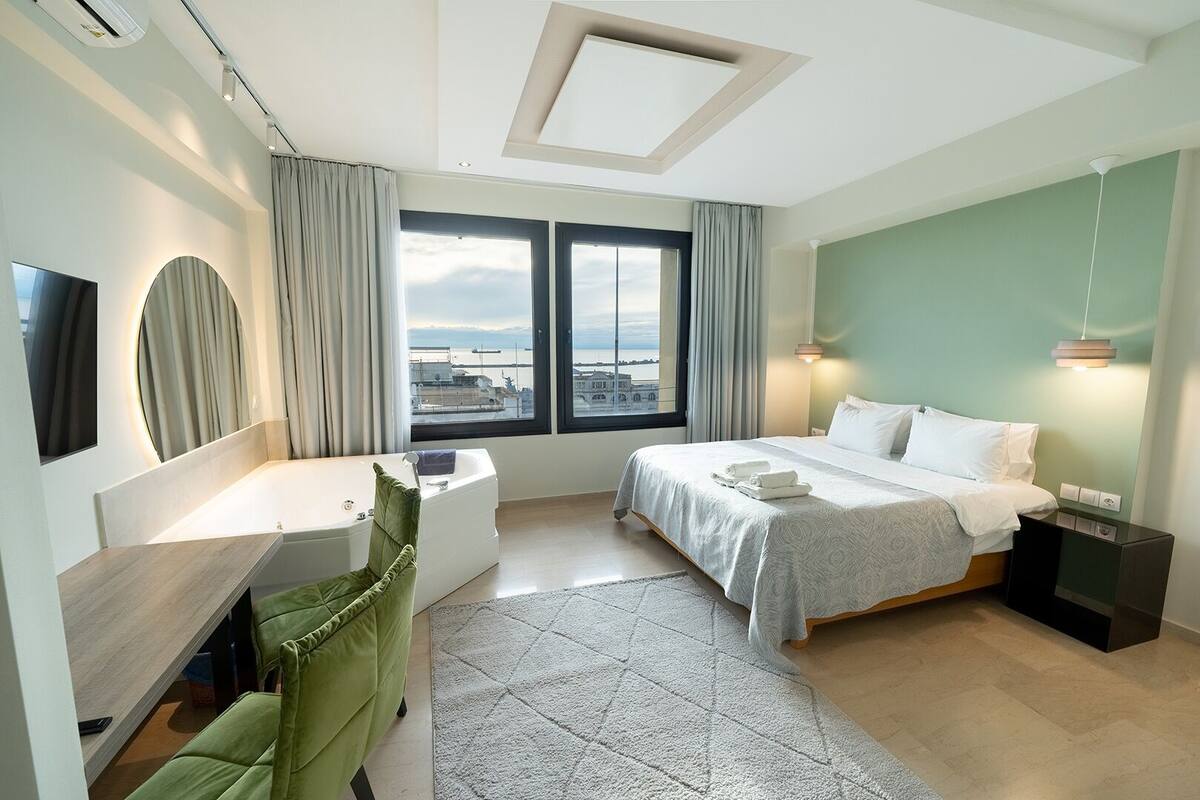 Een moderne hotelkamer met een bed, een groene fauteuil, een bureau en uitzicht op de stad door grote ramen.
