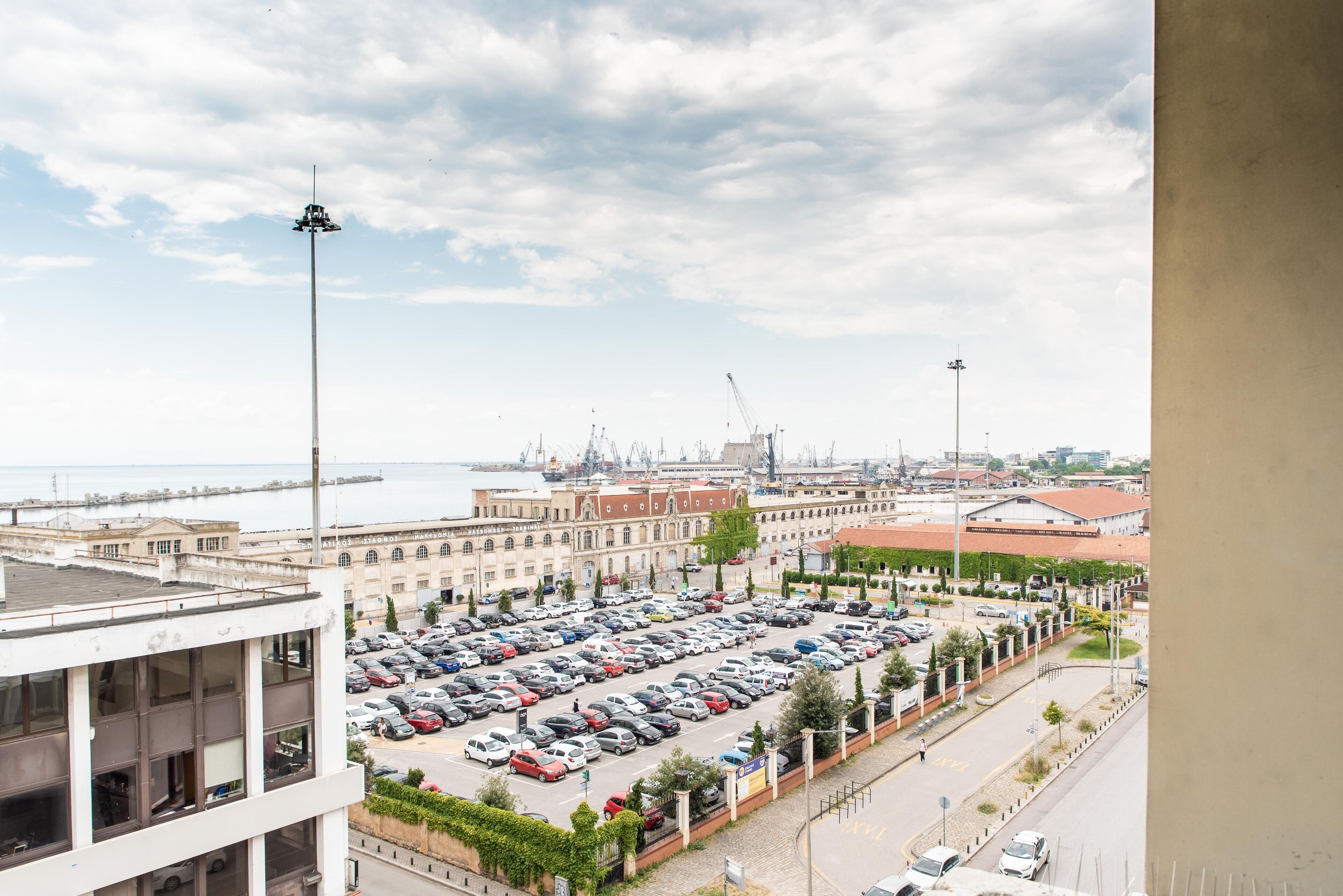 Un aparcamiento con varias filas de coches, un edificio con grandes ventanales y una vista al mar y al puerto en el fondo.