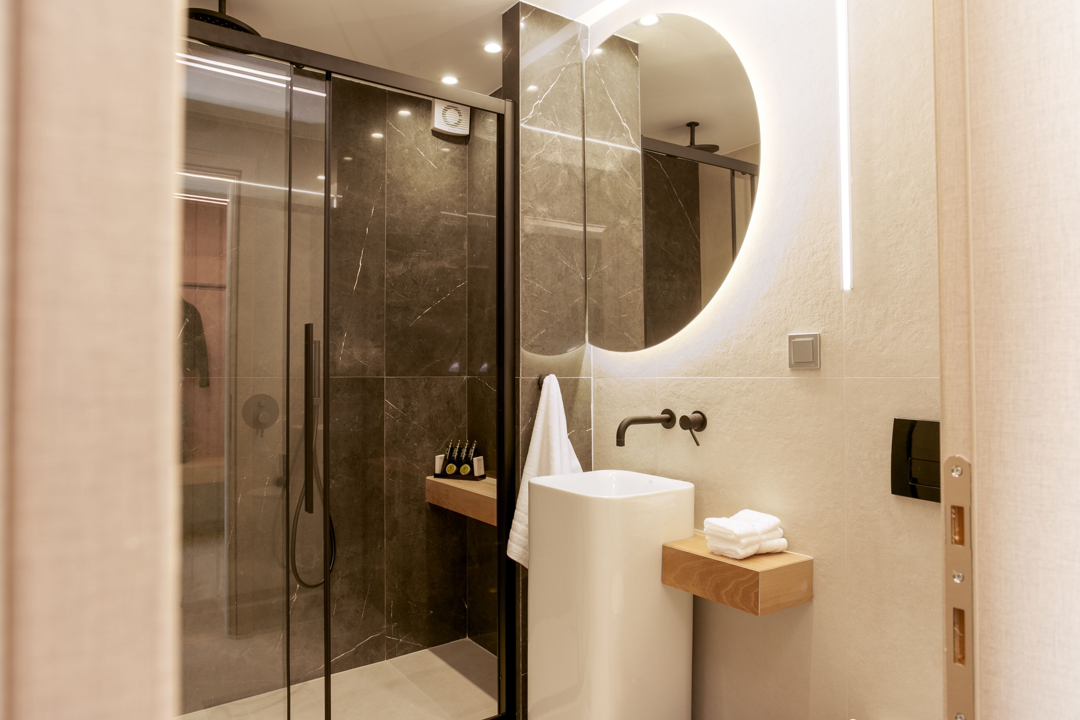 A modern bathroom with a glass-enclosed shower, a white sink, and a wooden shelf with toiletries.