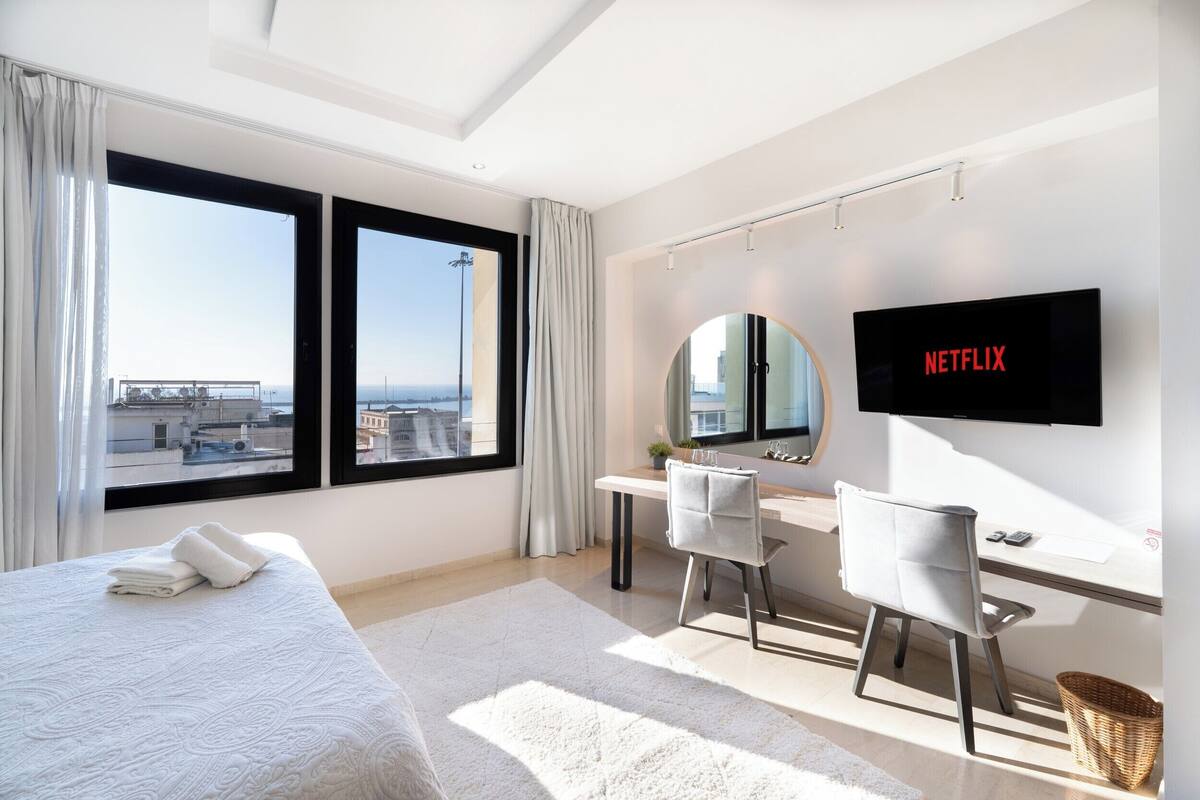 Een moderne hotelkamer met een groot bed, een bureau met stoel, een aan de muur bevestigde televisie en een stadszicht door grote ramen.