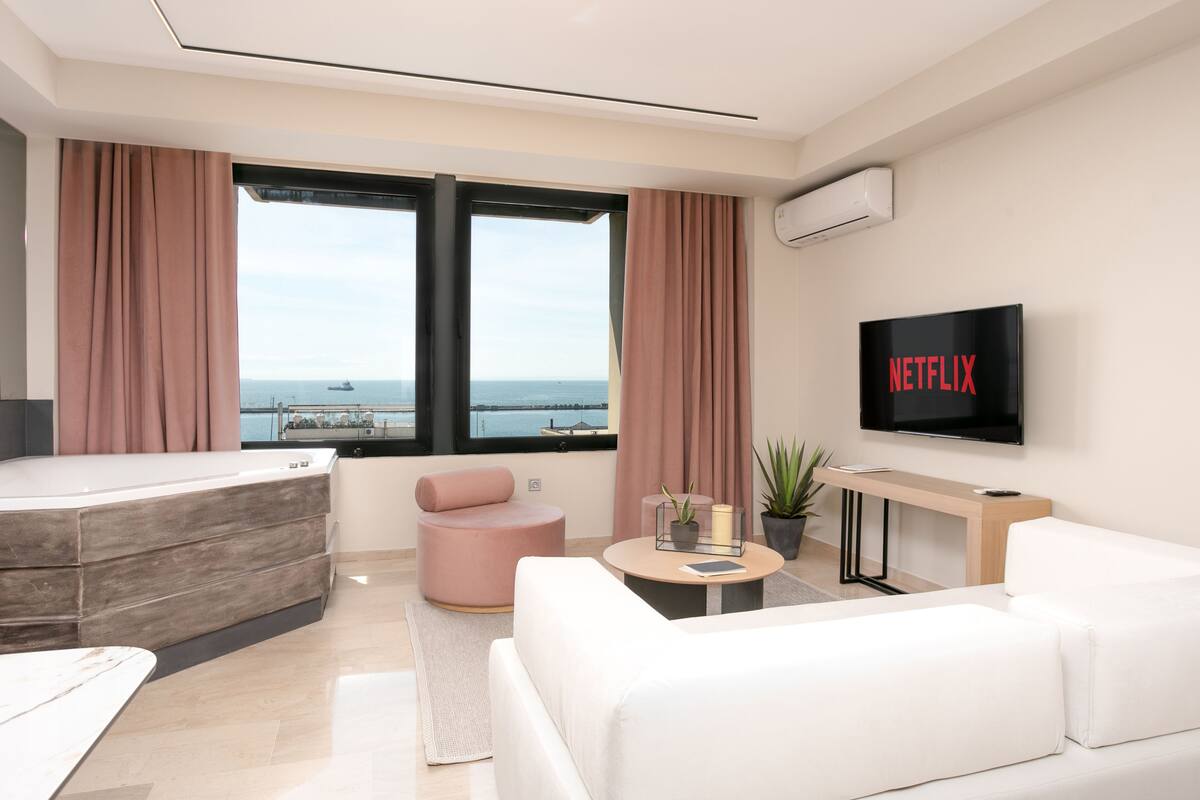 Een moderne hotelkamer met een groot raam, een flatscreen televisie, een bank, een stoel en een klein tafeltje met een plant.