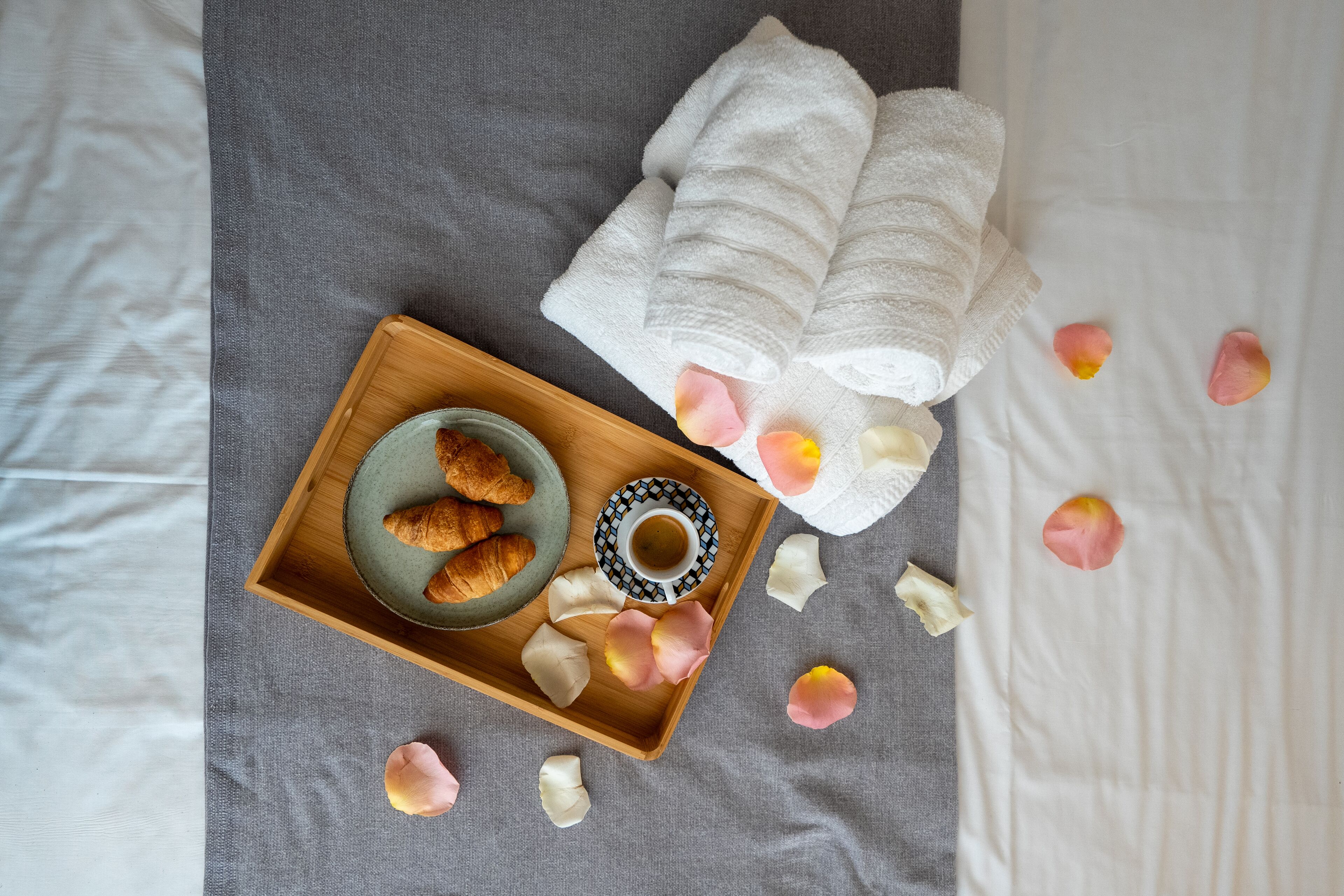 A wooden tray with croissants and a cup of coffee on a bed with white and gray bedding.