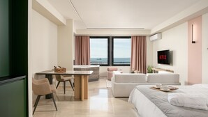 Een moderne hotelkamer met een groot bed, een zithoek met een tafel en stoelen, een aan de muur bevestigde televisie en uitzicht op zee door grote ramen.