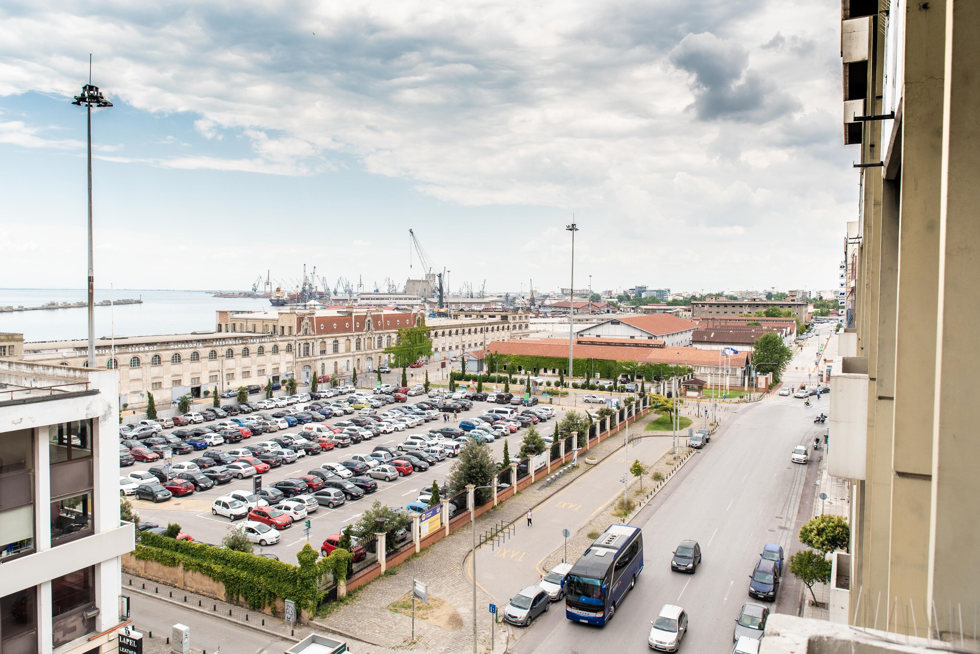 A parking lot with multiple rows of cars, a bus, and a view of a port with cranes in the background.