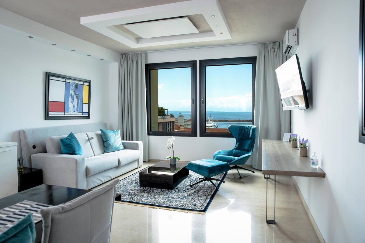 Een moderne woonkamer met een bank, fauteuil en salontafel, grote ramen met uitzicht en een flatscreen televisie aan de muur.